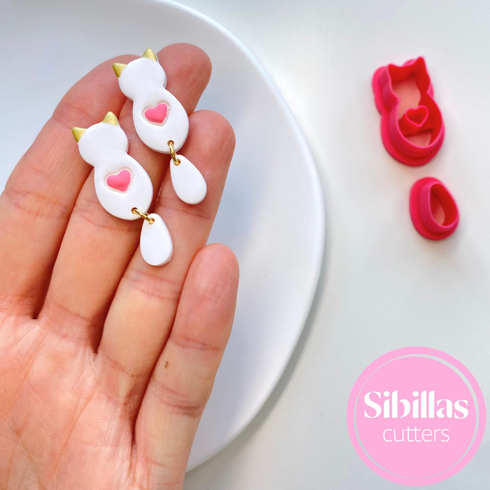 Hand holding cat-shaped cookie cutters with pink hearts on a white surface, branded 'Sibillas cutters'.