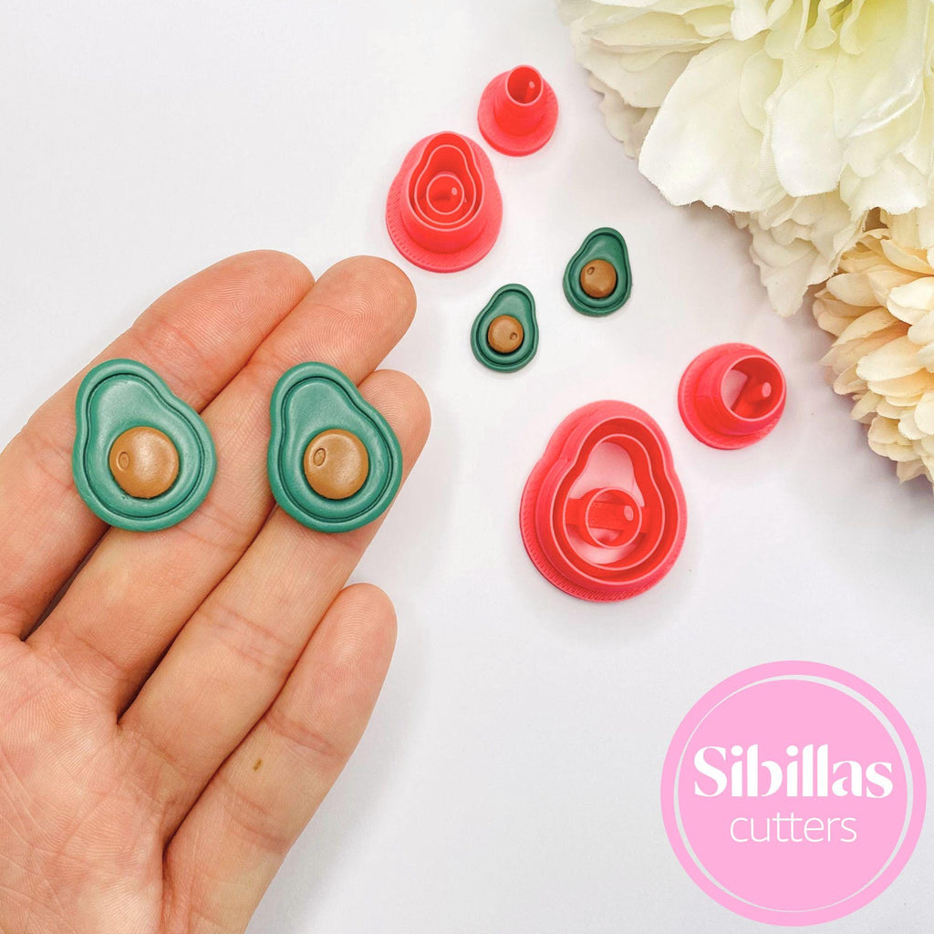 Avocado-shaped cookie cutters on a white surface with flowers, held by a hand.