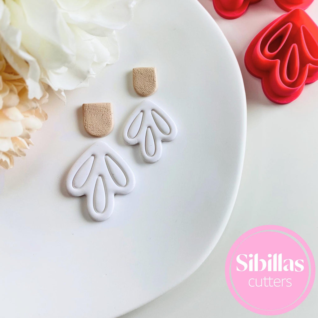 Heart-shaped cookie cutters on a white plate with flowers and additional cutters in the background, branded 'Sibillas Cutters'.