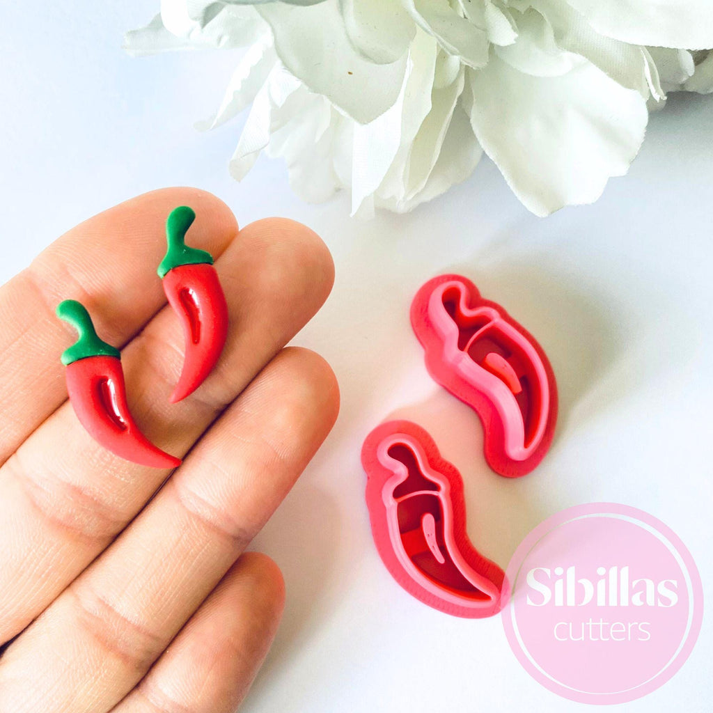Red chili pepper cookie cutters held by a hand with white flowers in the background, featuring the brand 'Sibillas Cutters'.