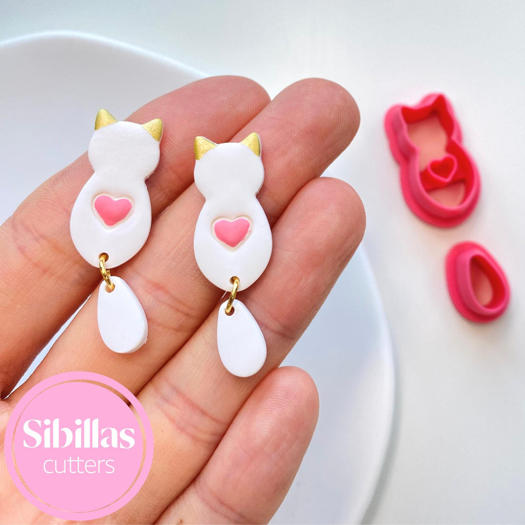 White cat-shaped cookie cutters with pink hearts held in a hand, branded 'Sibillas Cutters'.