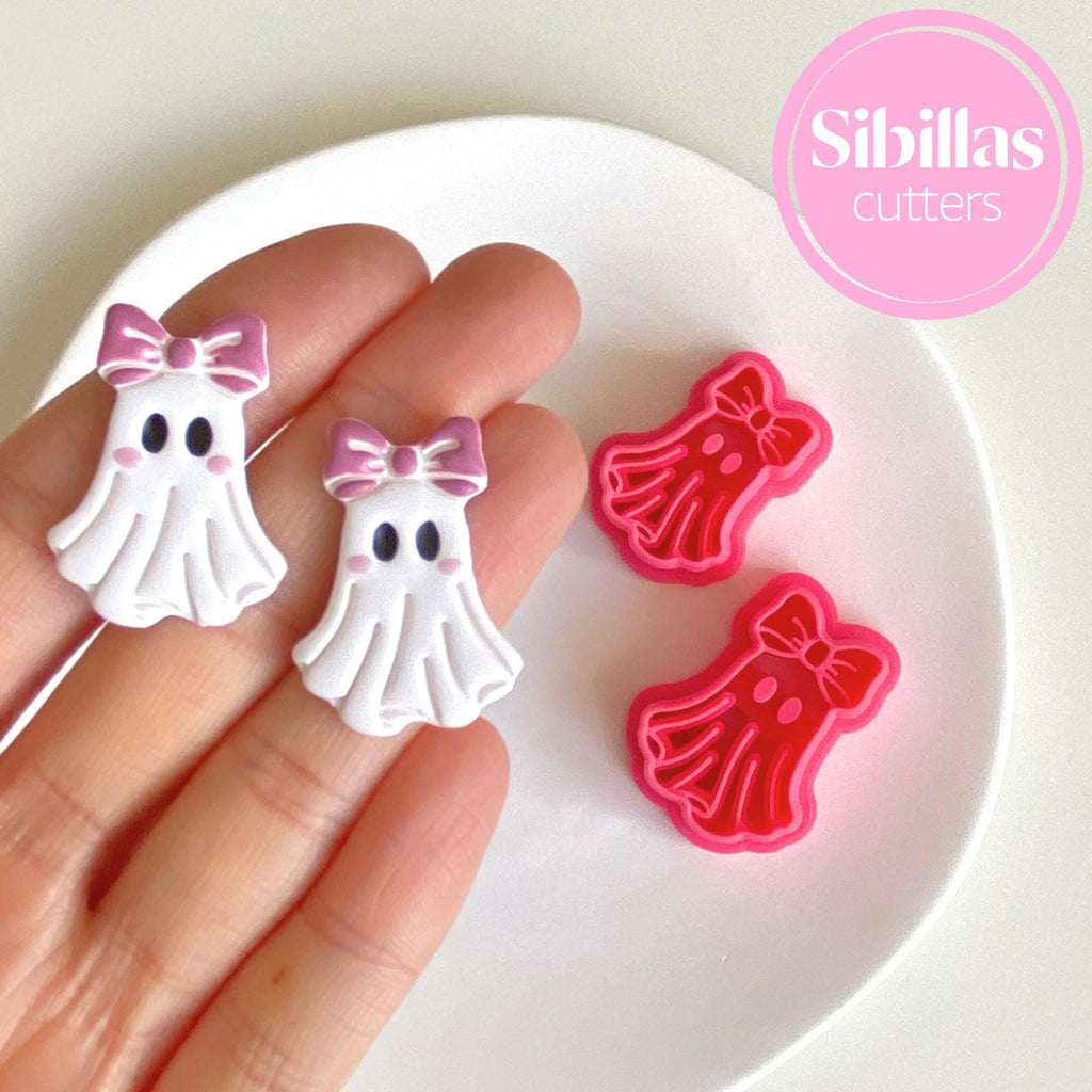 Ghost-shaped cookie cutters with pink bows held by a hand on a white plate, branded 'Sibillas'.