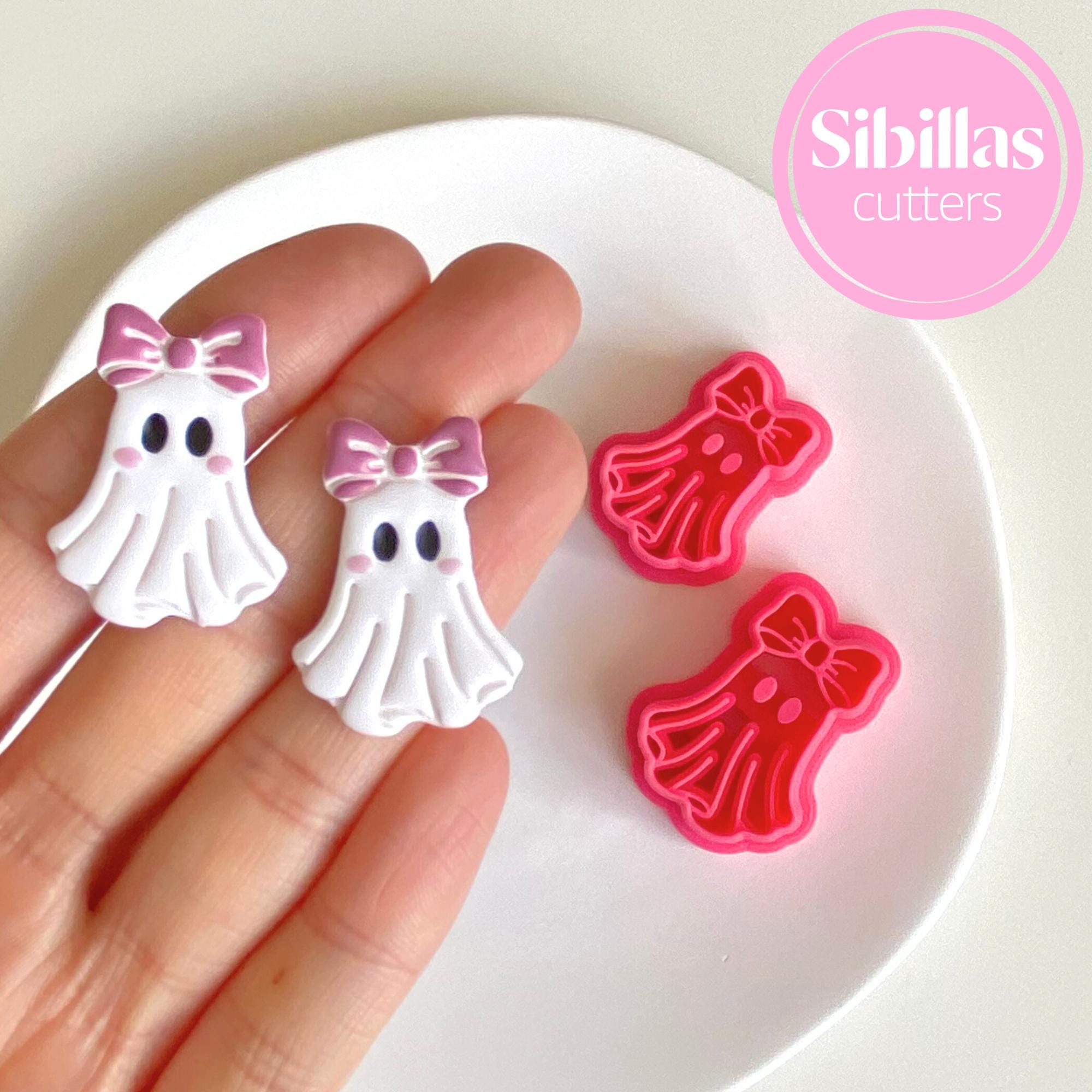 Ghost-shaped cookie cutters with pink bows held by a hand on a white plate, branded 'Sibillas'.
