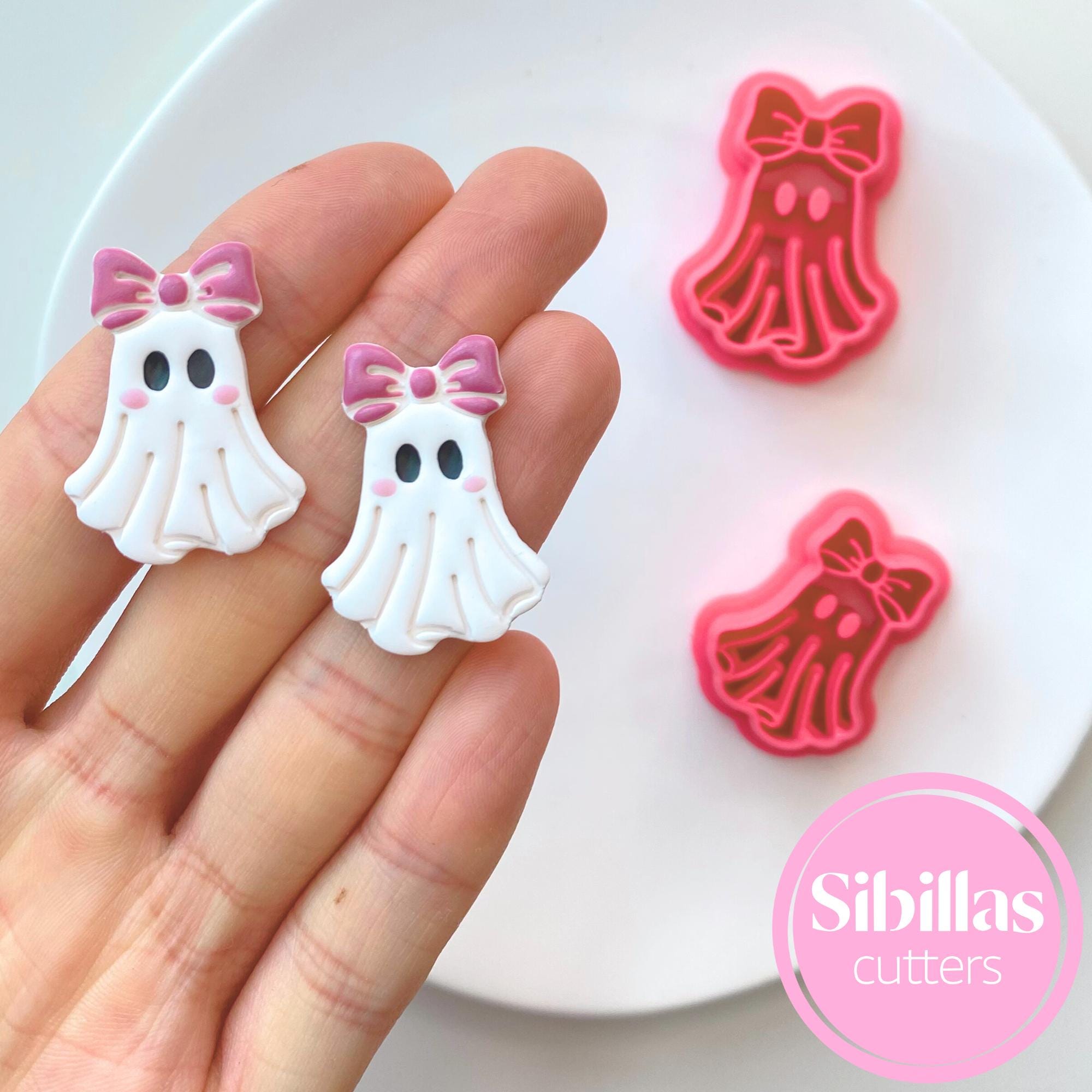 White ghost-shaped cookie cutters with pink bows held by a hand, on a white plate with 'Sibillas cutters' branding.