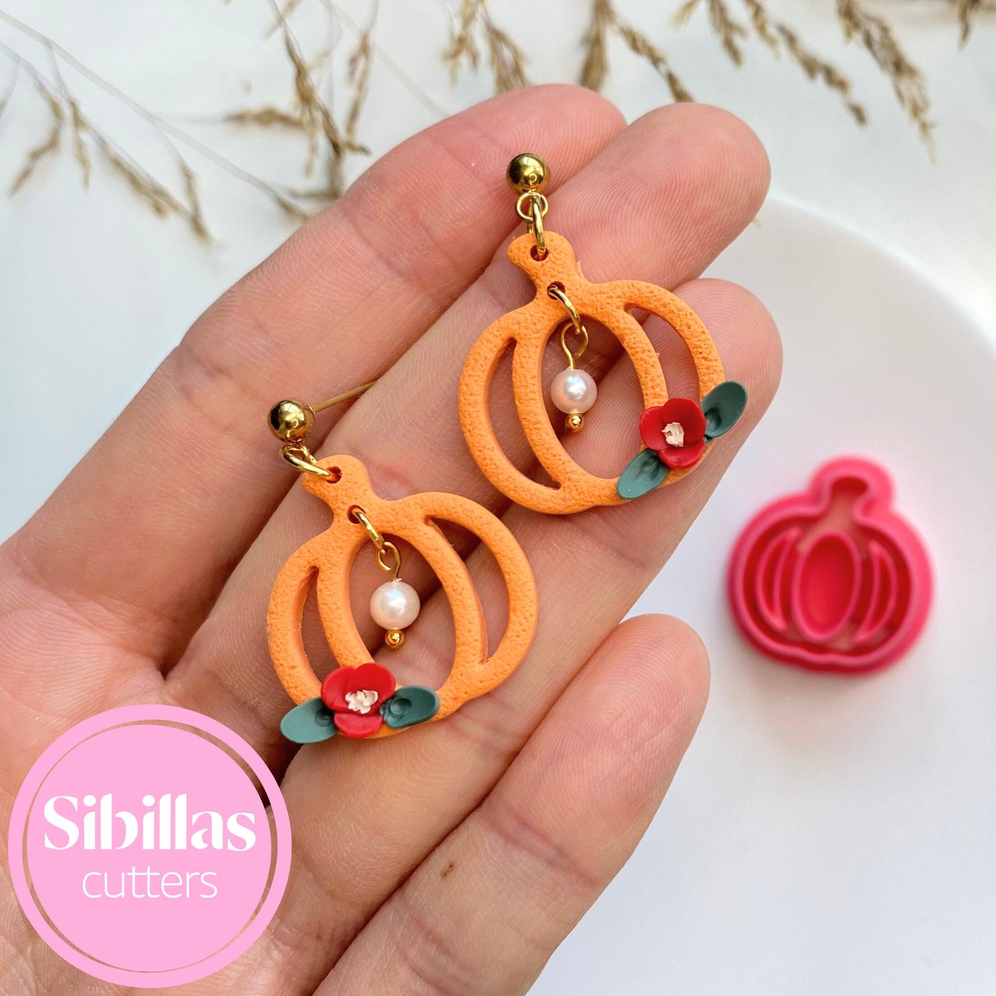 Orange pumpkin-shaped earrings with floral details held in a hand, with 'Sibillas Cutters' logo.