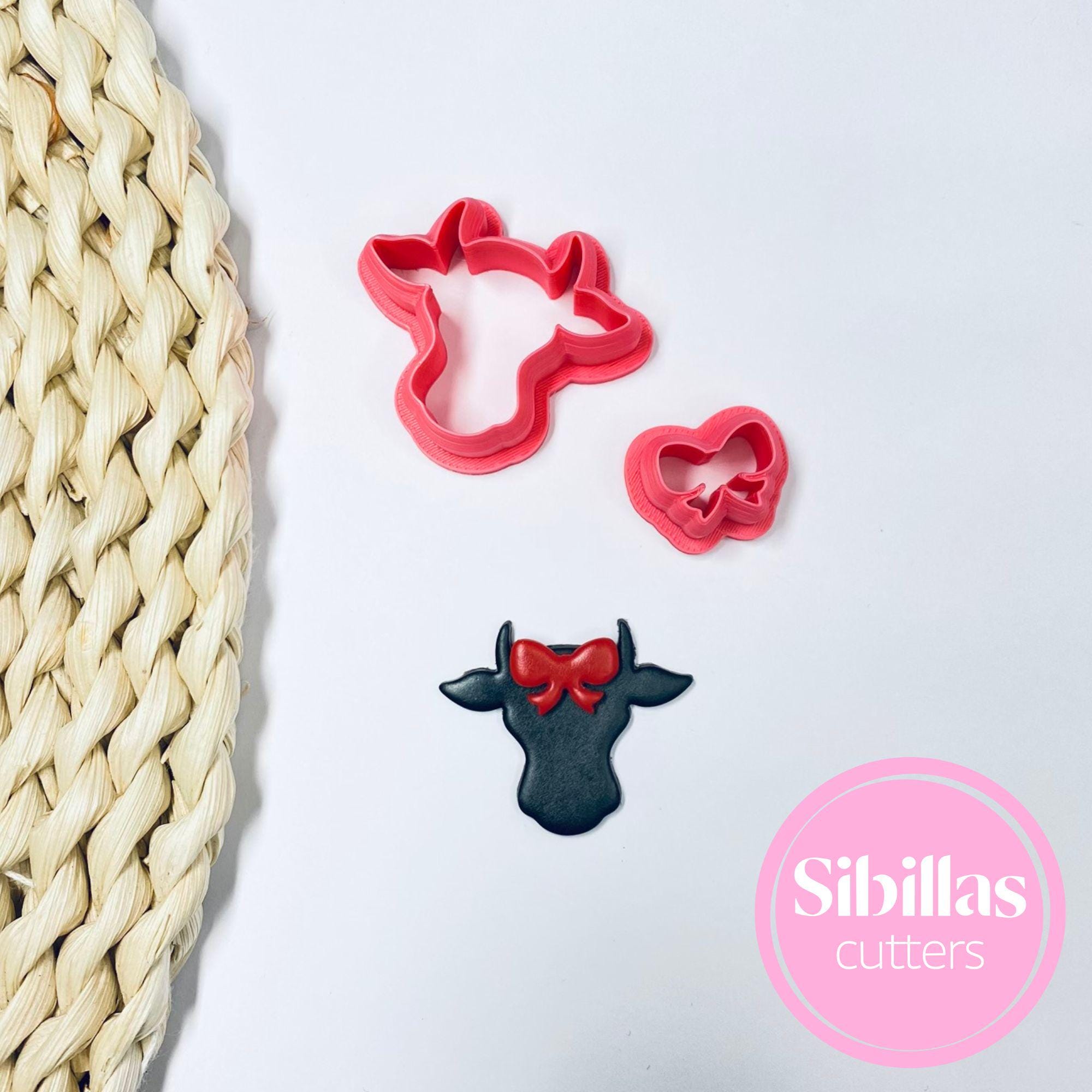 Red cow-shaped cookie cutter with a black cow-shaped cookie on a white surface, next to a woven mat. Sibillas Cutters logo visible.