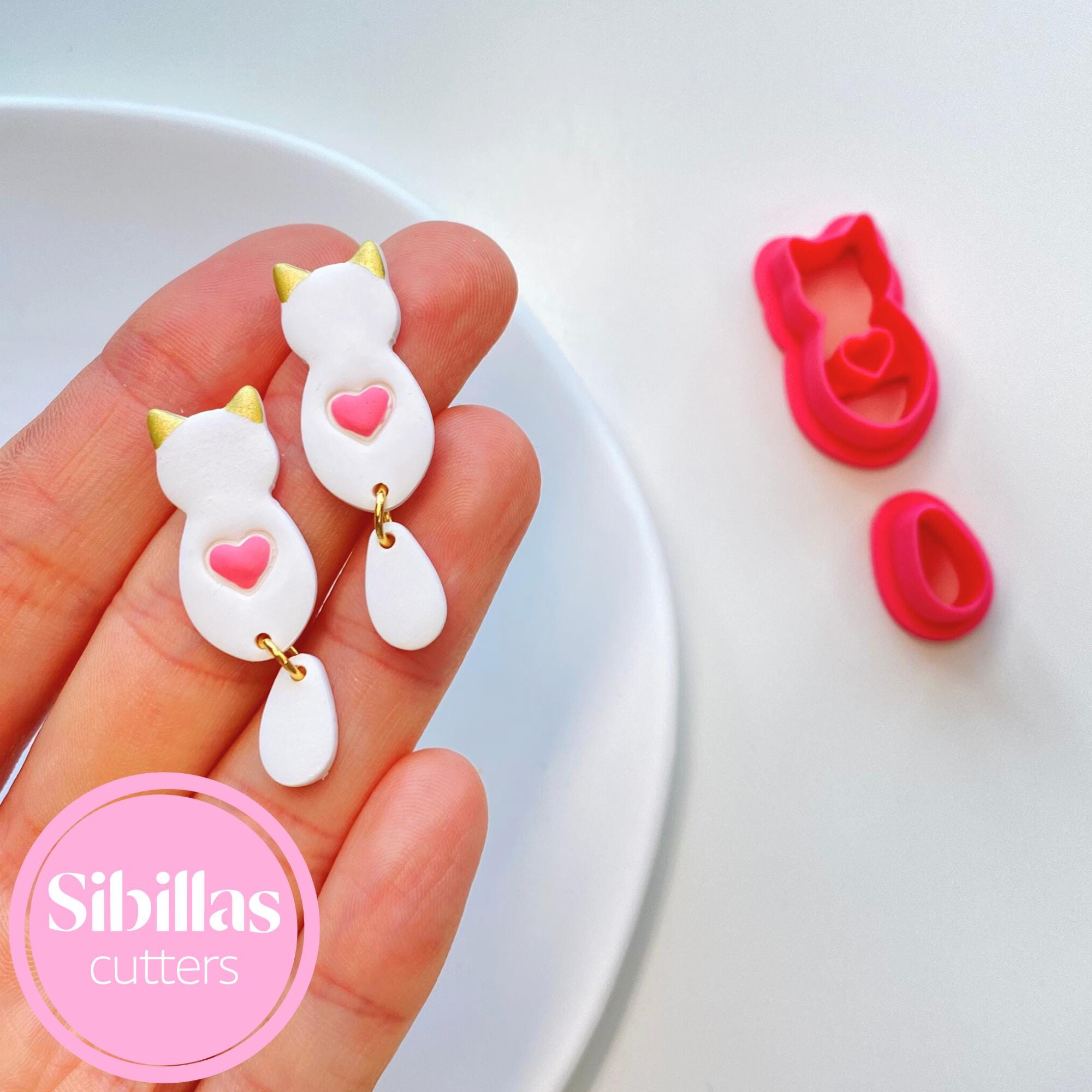 Hand holding a white cat-shaped cookie cutter with pink heart and yellow accents, next to red heart-shaped cookie cutters on a white surface.