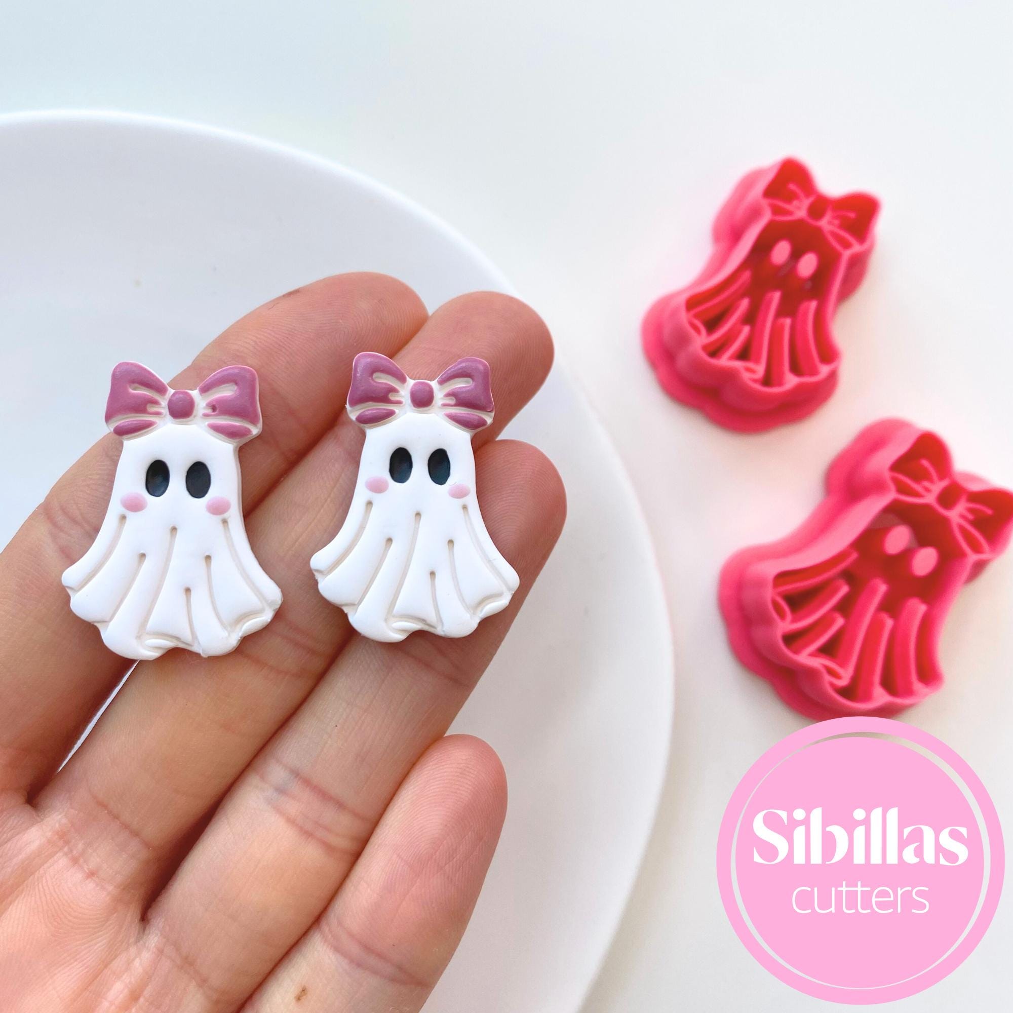 White ghost-shaped cookie cutters with pink bows held by a hand, next to red ghost-shaped cookie cutters on a white background.