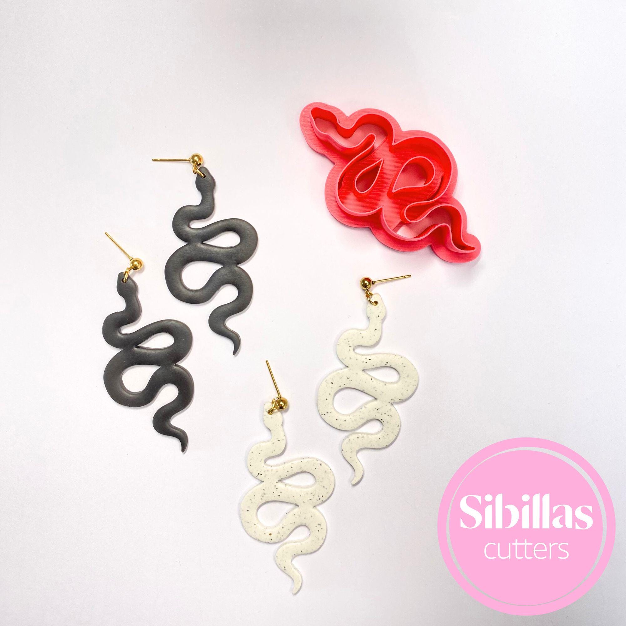 Snake-shaped earrings in gray, white, and red with a cookie cutter on a light gray background. Sibillas Cutters logo visible.