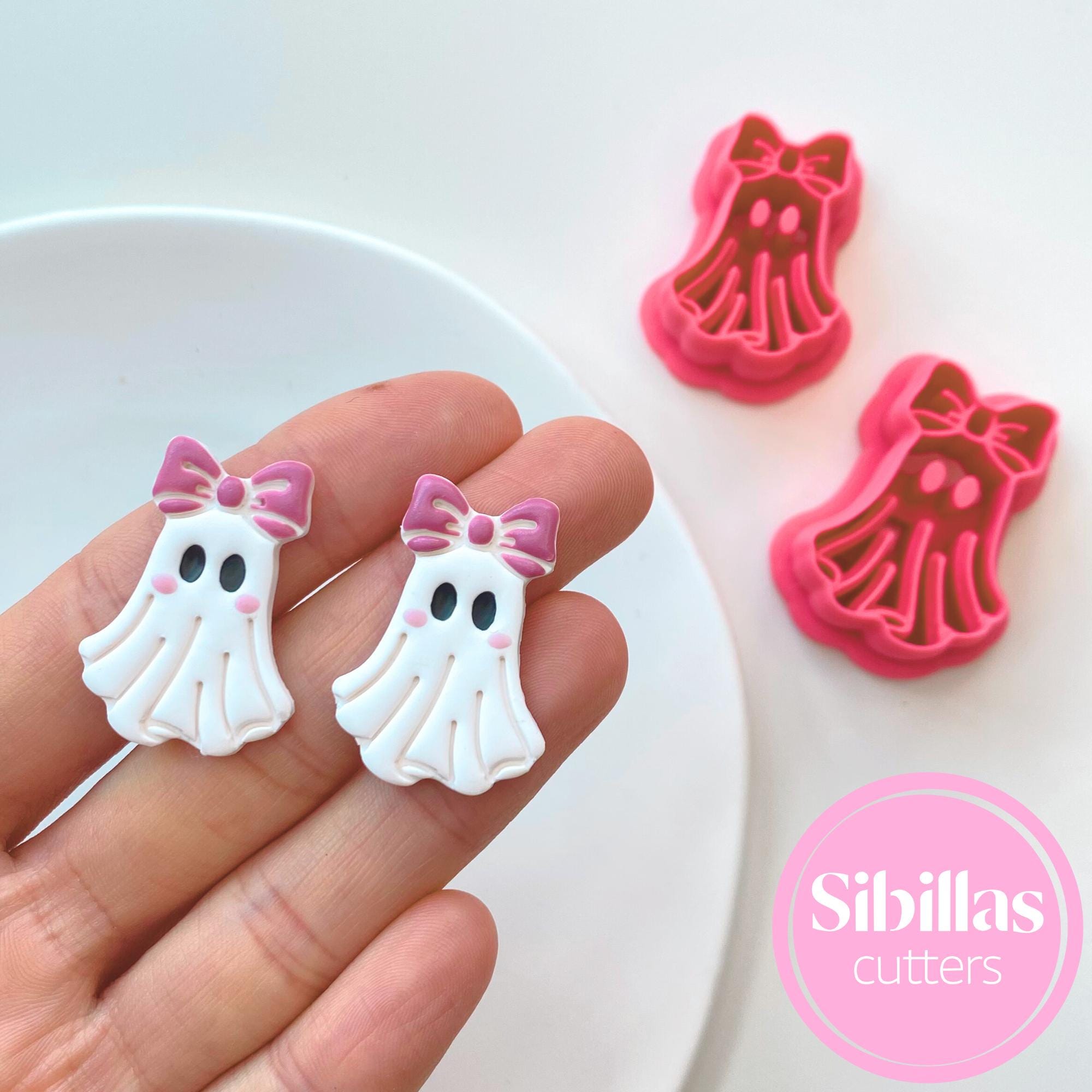 White ghost-shaped cookie cutters with pink bows held by a hand, next to pink and red dress-shaped cutters on a white background.