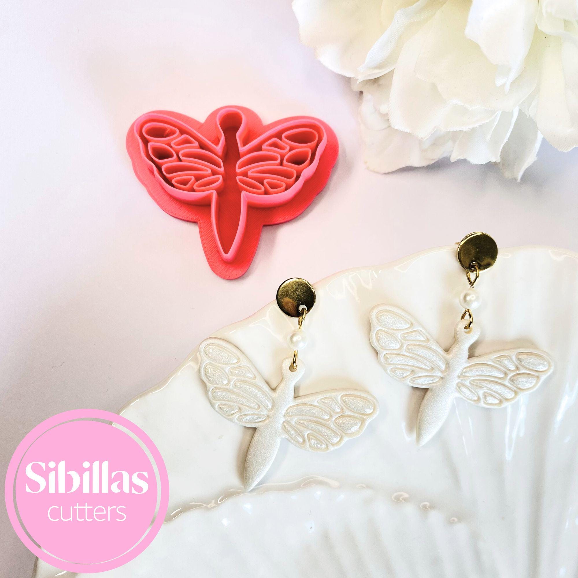 Butterfly-shaped cookie cutter with butterfly earrings on a pink surface, featuring the brand 'Sibillas Cutters'.