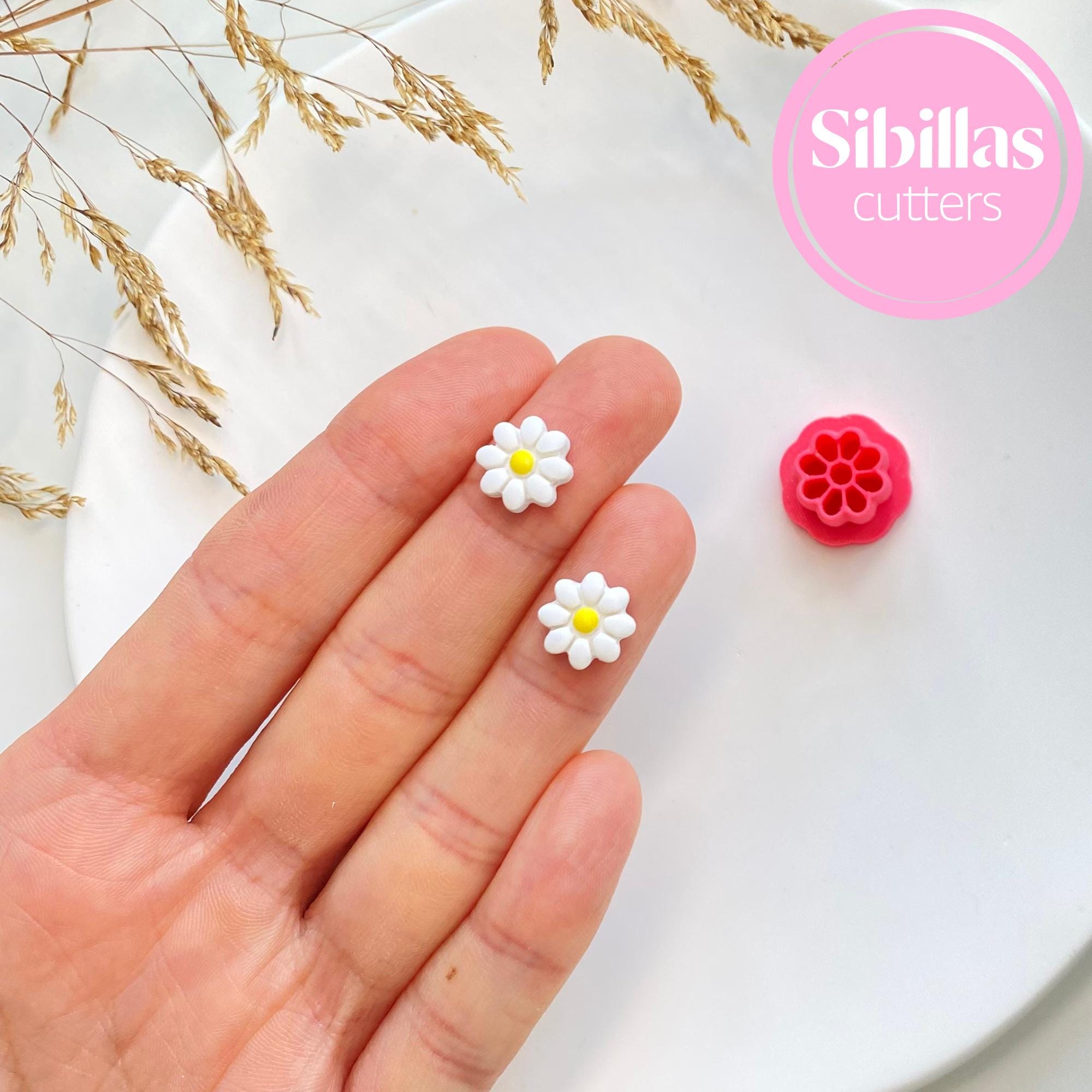 Hand holding two small flower-shaped cutters with a pink 'Sibillas cutters' logo in the corner.