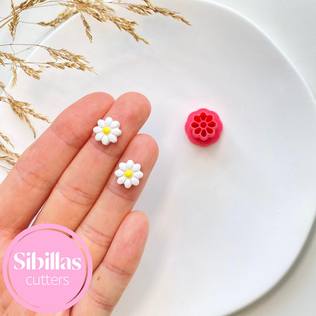 Hand holding two flower-shaped cutters with a white plate and decorative grasses in the background, featuring the brand 'Sibillas cutters'.