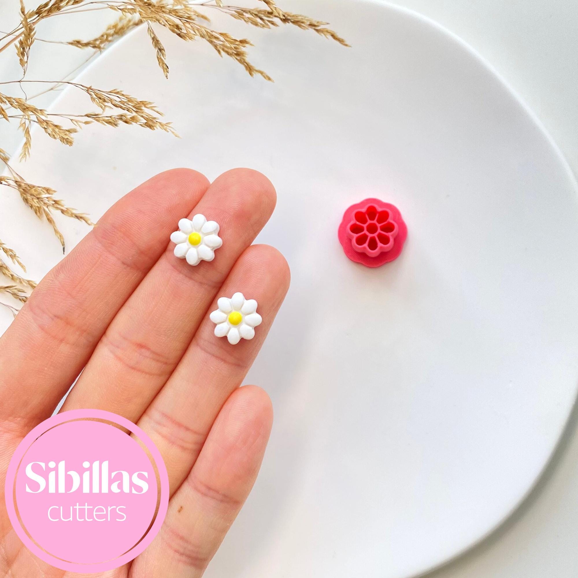 Hand holding two flower-shaped cutters with a white plate and decorative grasses in the background, featuring the brand 'Sibillas cutters'.