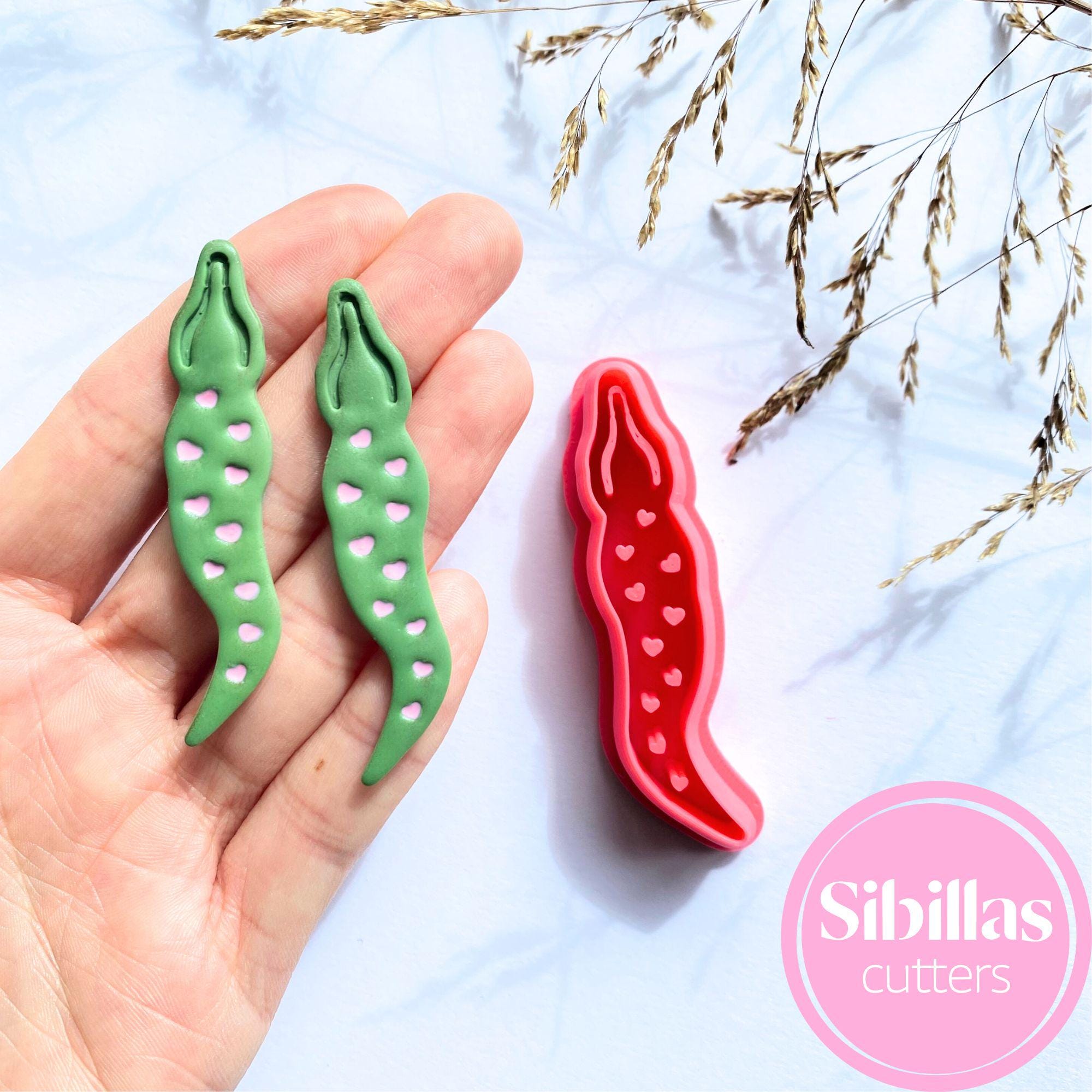 Green and red cookie cutters shaped like peas and beans held by a hand on a light blue background with 'Sibillas Cutters' branding.