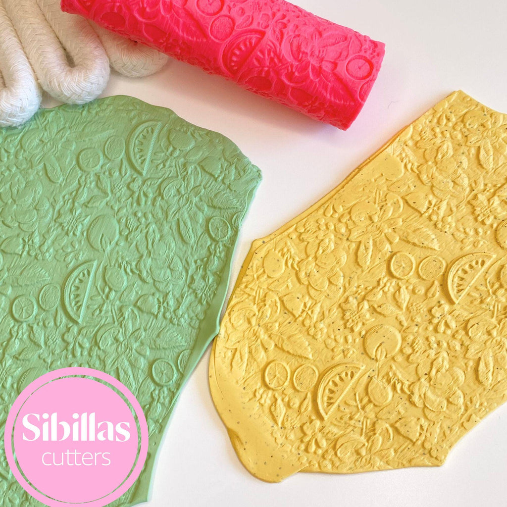 Two embossed cookie cutters, one green and one gold, with a pink rolling pin on a white background. Sibillas Cutters logo is visible.