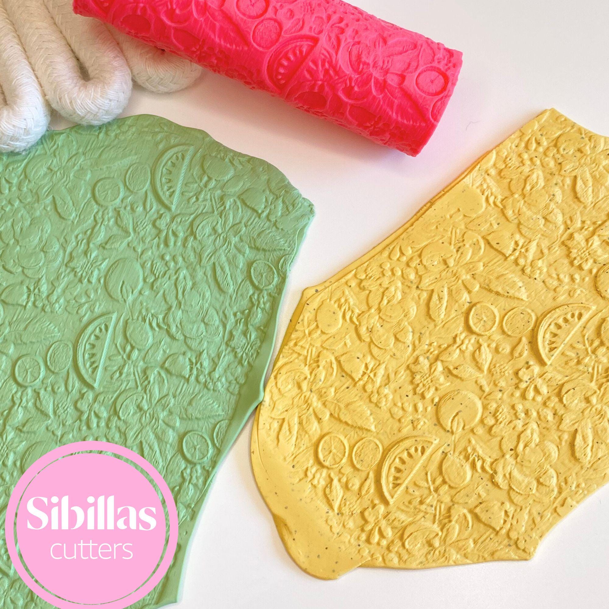 Two embossed cookie cutters, one green and one gold, with a pink rolling pin on a white background. Sibillas Cutters logo is visible.