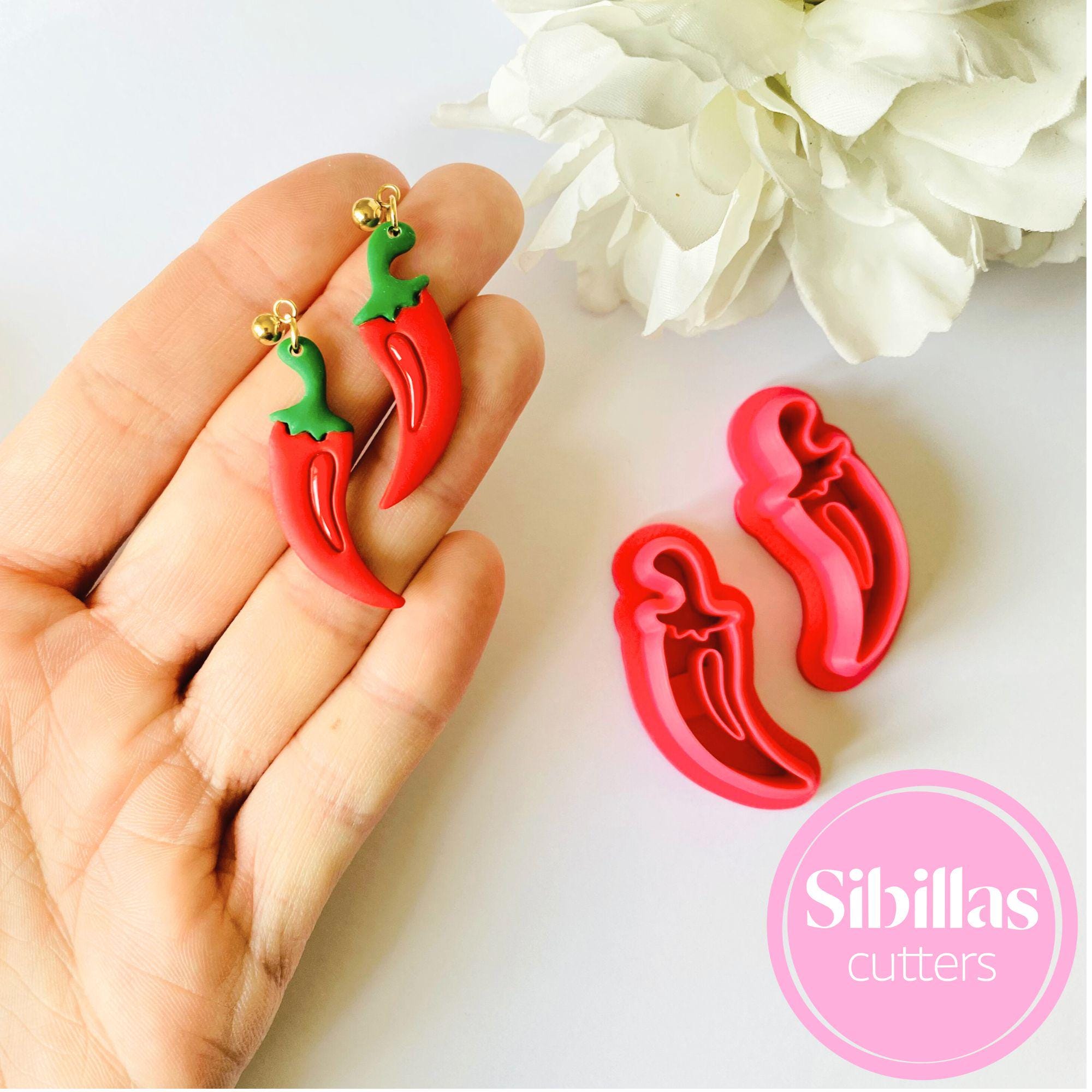 Red chili pepper-shaped cookie cutters held in a hand with white flowers in the background, branded 'Sibillas cutters'.