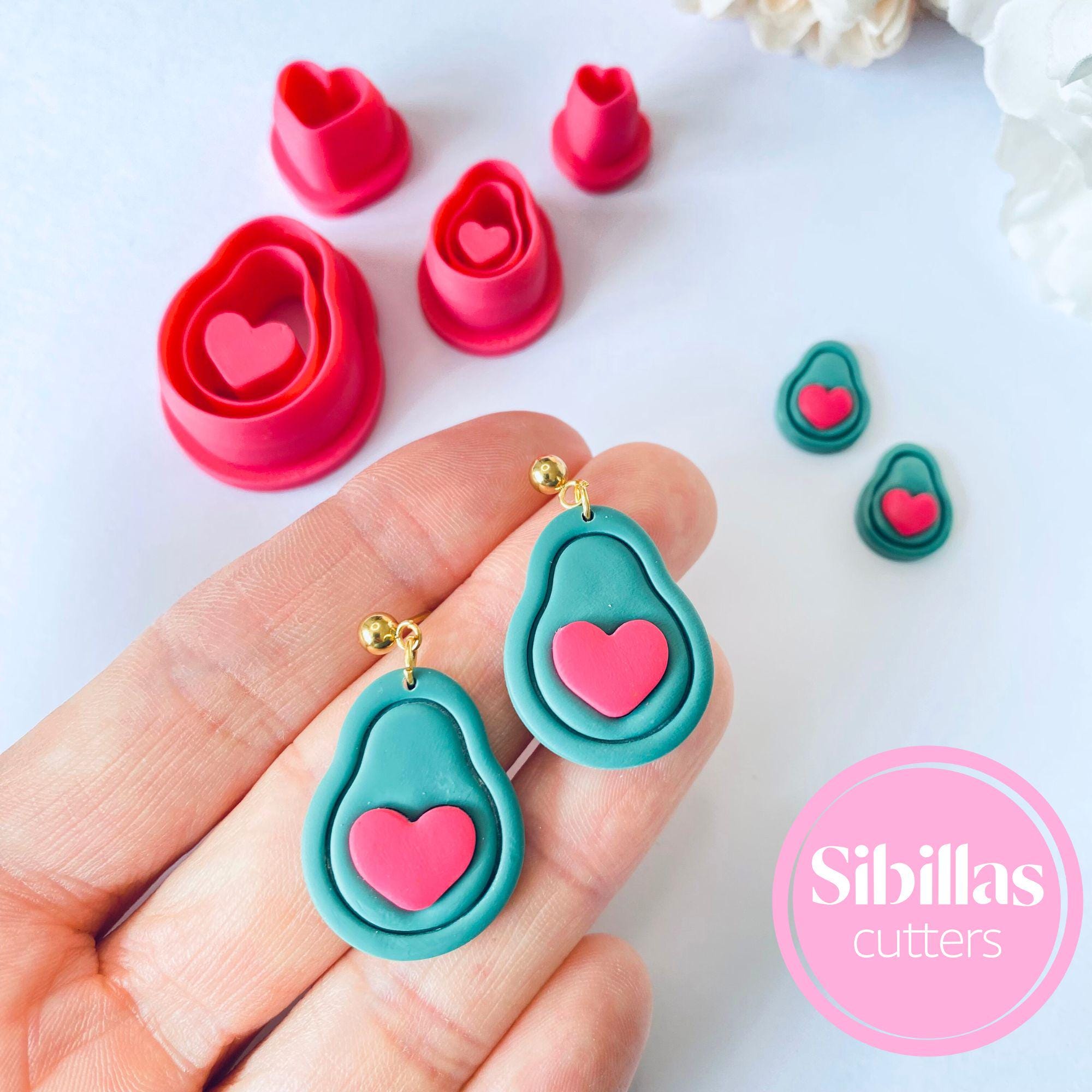 Teal earrings with pink hearts held in a hand, surrounded by heart-shaped cookie cutters on a white background.